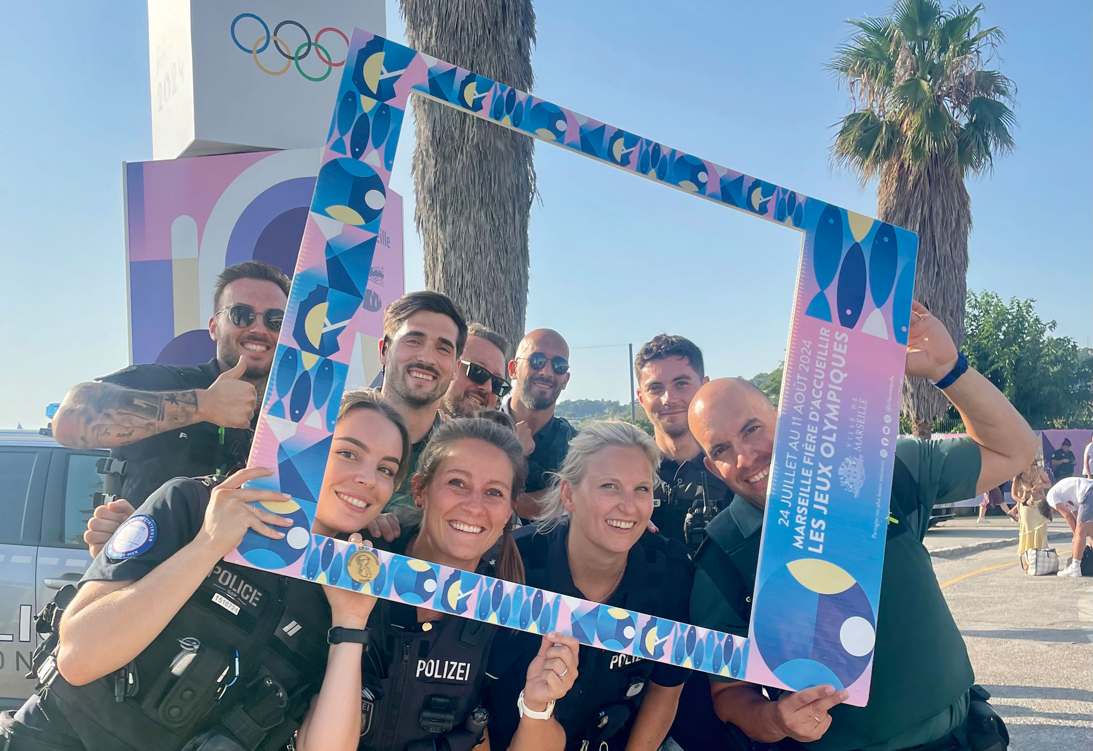 Deutsche Polizistin Kristina Kilb umringt von acht französischen Polizistinnen und Polizisten bei den Olympischen Spielen in Paris. Im Hintergrund sieht man eine Palme und die olympischen Ringe. Gemeinsam halten sie einen Fotorahmen, durch den sie durchschauen