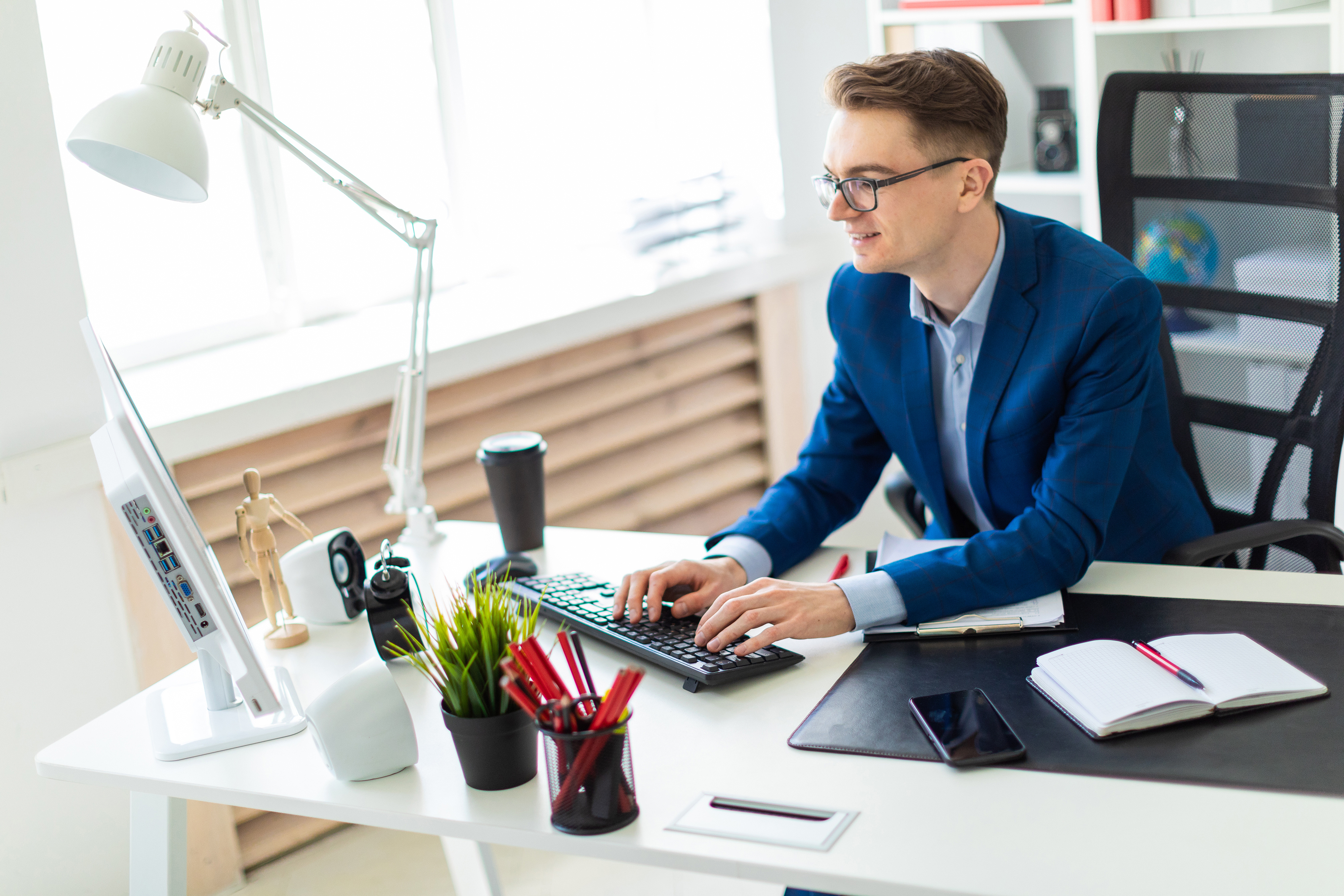 Junger Mann mit Brille arbeitet an einem modernen Schreibtisch. Er ist mit Hemd und Blazer bekleidet, vor ihm auf dem Tisch stehen ein Monitor, kleine Lautsprecherboxen, ein Wecker, eine Pflanze und ein Stifthalter. Handy, Kalender und ein Kaffeebecher. Die Einrichtung des Büros ist hell und freundlich. 