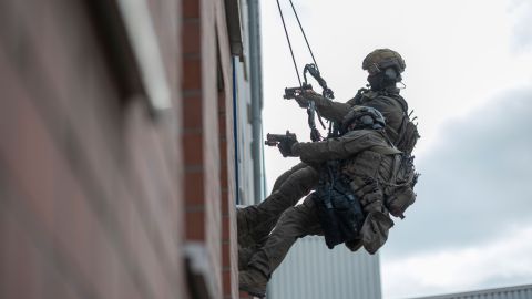 Two SEK officers from the climbing team abseil down the outside of a house wall into a window. They point their weapons into the room.