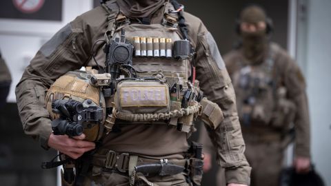 Im Vordergrund ist ein SEK-Beamter in einem Ausschnitt vom Hals bis zu den Oberschenkeln zu sehen. Er trägt eine Uniform und zahlreiche Ausstattungsgegenstände, sowie mehrere Waffen. Im Hintergrund ist ein weiterer SEK-Beamter zu sehen.